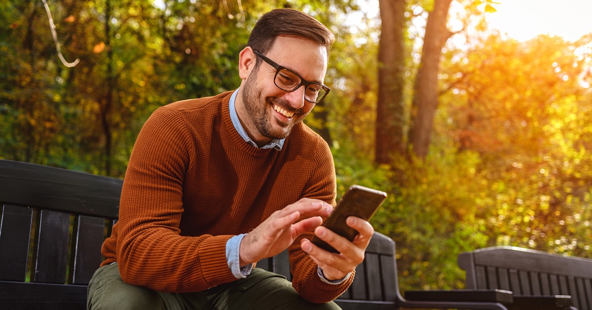 A man sits outside with autumn leaves behind him, smiling at his phone